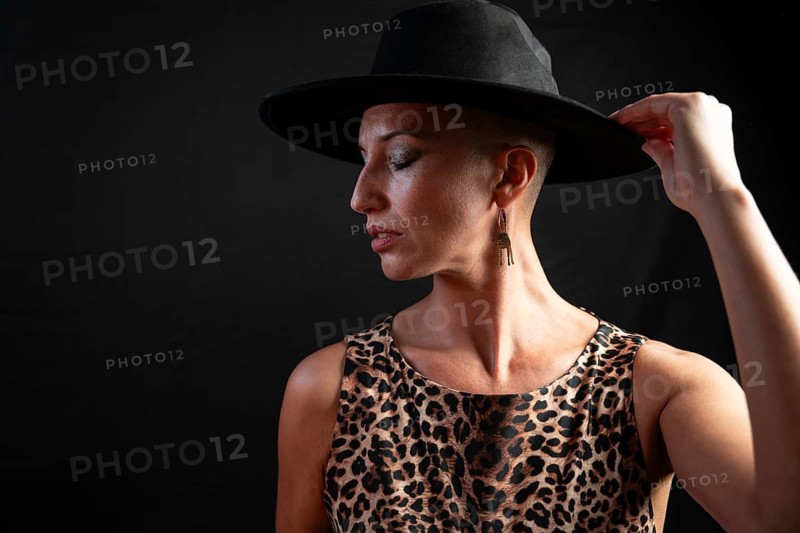 Portrait of a bald woman wearing a leopard print dress and black hat. She adjusts her hat, showcasing distinctive dangling earrings, against a dark backdrop