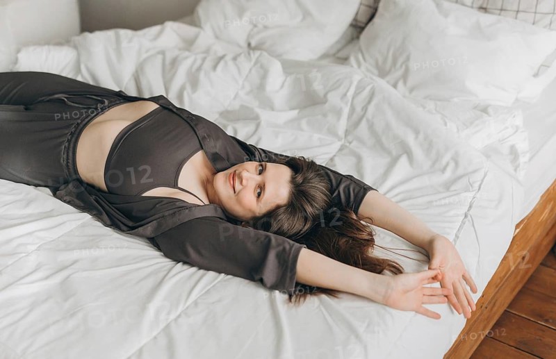 A cheerful young woman lies on a white bed with her arms stretched out, smiling and looking relaxed. The cozy setting and natural light create a warm and inviting atmosphere