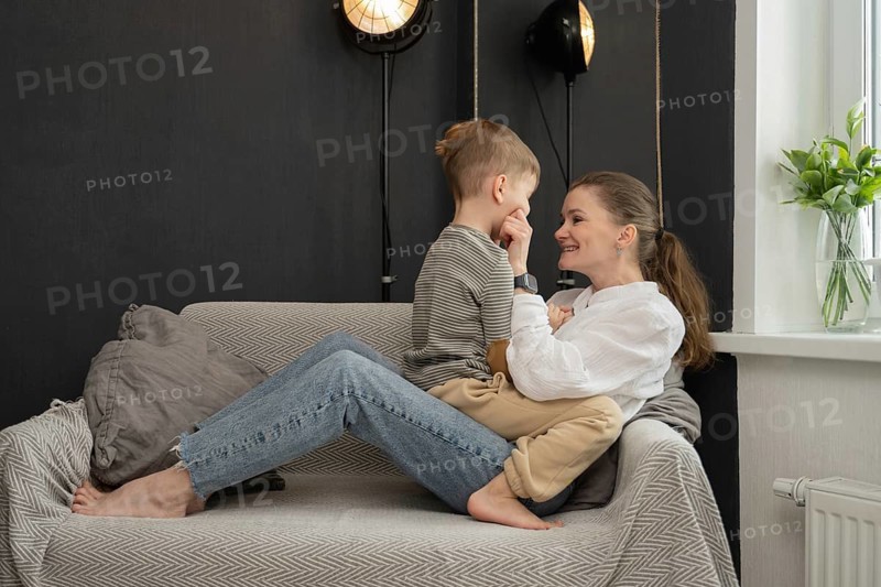 A tender interaction between a mother and her child, sitting on a comfortable sofa. The setting is a cozy living room with soft lighting and a vase of fresh green leaves by the window