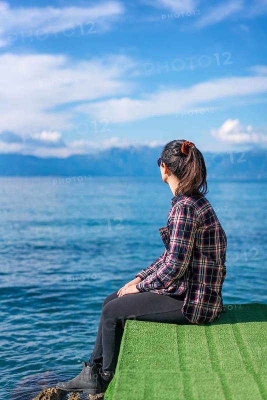 A woman wearing plaid sits on a green platform, gazing at the tranquil ocean under a clear blue sky in Greece. The serene water and distant mountains create a peaceful scene