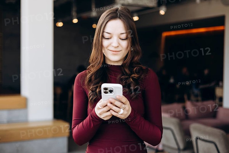 A young female student with long hair stands indoors, focused on her smartphone. She wears a burgundy dress and smiles gently, immersed in her digital world