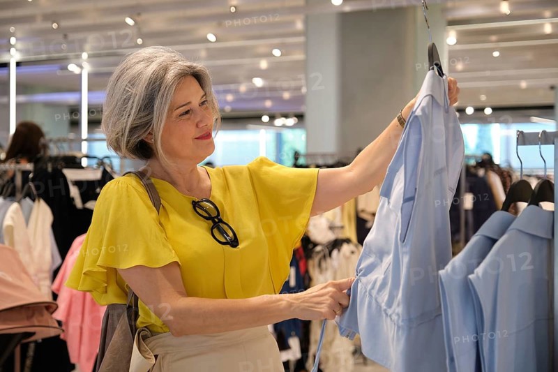 A mature woman with gray hair browses a clothes rack in a store, holding a blue shirt and wearing a yellow blouse. She appears content and focused on her shopping experience