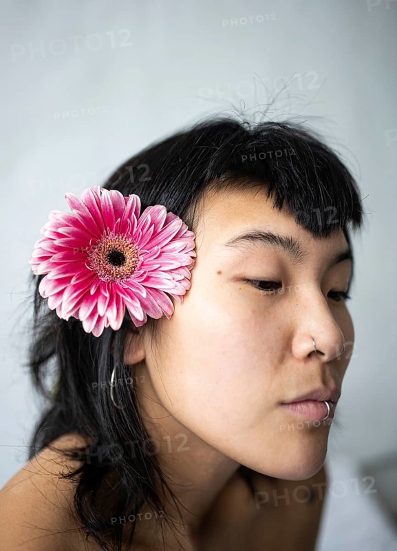 A serene portrait of an Asian woman with a pink flower tucked behind her ear. Her calm expression and the soft light create a sense of tranquility and grace