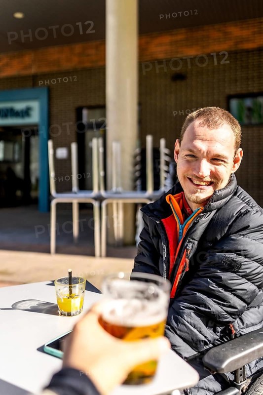 Young disabled man smiling and enjoying a beer with a friend at an outdoor cafe, promoting inclusion and social connection