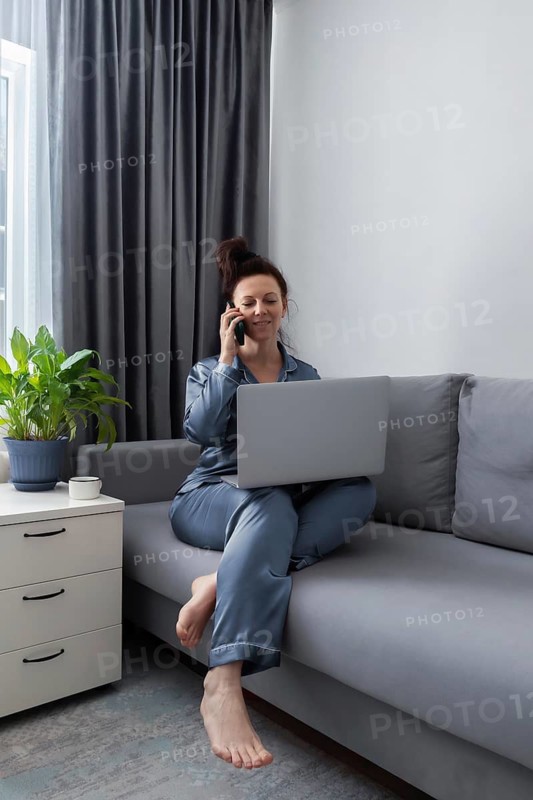 A woman in pajamas multitasks by using a phone and laptop on a gray couch. A potted plant and coffee cup add a cozy touch to the home office setting