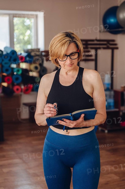 A female personal trainer in gym attire reviews her planner, organizing client workouts. She stands in a gym environment, exemplifying fitness and professional dedication