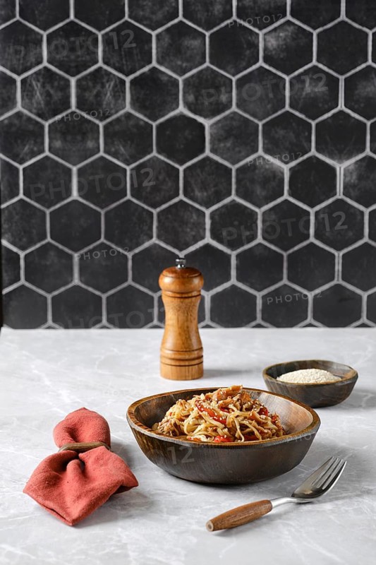 A bowl of spaghetti with pulled beef topped with fresh herbs and spices on a marble countertop, accompanied by a pepper grinder and a small dish of grated cheese
