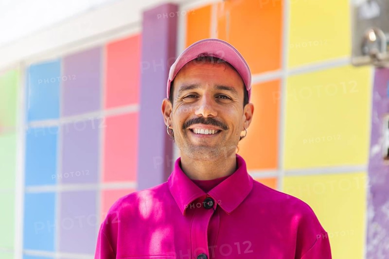 A Spanish man with a mustache models vibrant pink clothing in Quito, Ecuador. He displays a joyful urban lifestyle with a colorful background, reflecting casual fashion and themes of self-expression and diversity associated with LGTBIQ+