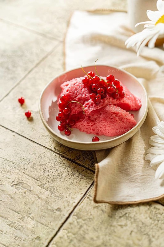 From above homemade pink berry mousse served in a bowl, topped with a cluster of fresh red currants, accompanied by flowers on a tiled table