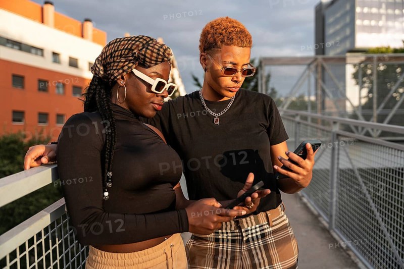 An LGBT multiethnic couple stands on a bridge, holding phones and enjoying each other's company. They're both wearing sunglasses, with urban buildings in the background