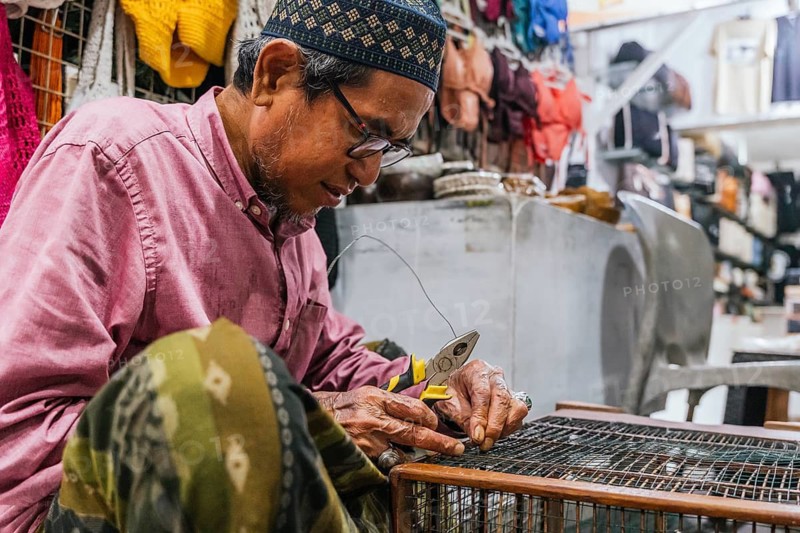 A balinese craftsman repairs a bird cage using pliers in a vibrant workshop. Surrounded by colorful fabrics and tools, showcasing traditional craftsmanship and dedication
