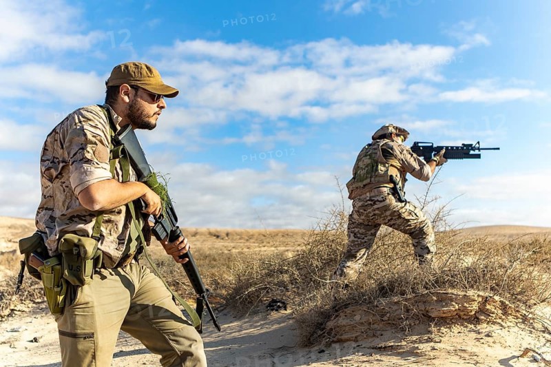 Two armed soldiers in desert terrain, wearing tactical gear and camouflage. Engaged in military training under a clear blue sky. Focus on readiness and teamwork