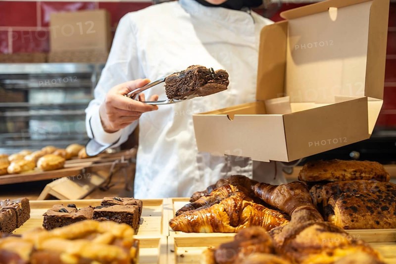 Baker wearing a chef uniform standing behind a counter, using tongs to carefully place a chocolate brownie into a cardboard takeaway box, with various pastries displayed on wooden trays
