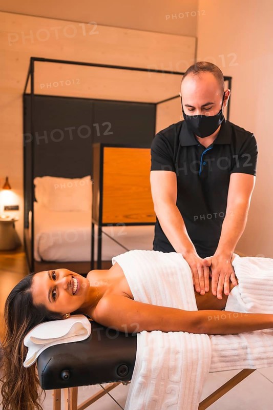 Home massage, a young man performing a massage at the client's home, vertical photo. With a face mask in the coronavirus pandemic