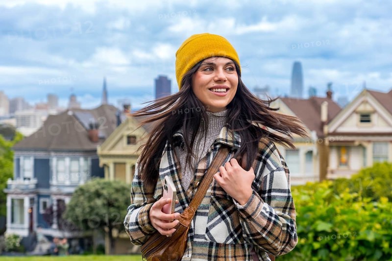 Young woman smiling while exploring san francisco, strolling past colorful victorian painted ladies with the skyline in the background on a bright city day
