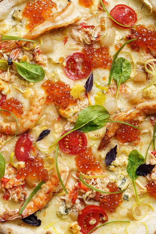 Close up view of a luxurious pizza adorned with succulent shrimp, crab meat, scallops, red caviar, and a variety of cheeses including mozzarella and blue cheese, complemented by slices of fresh cherry tomatoes and vibrant green spinach leaves