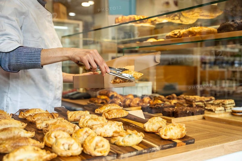 Baker's arms visible, picking up a hot empanada with tongs from the display counter, placing it into a brown takeaway box, with many other pastries and fresh food items surrounding the scene