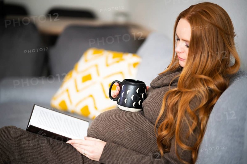 A pregnant woman with long red hair sits comfortably on a couch, reading a book and holding a mug. She exudes relaxation, embodying the calmness of maternity