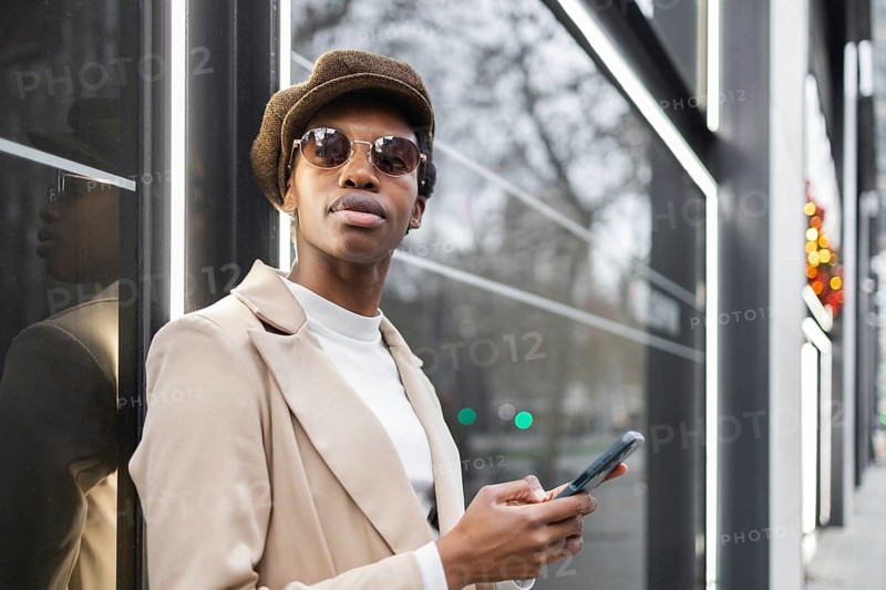 Chic black woman in an urban setting, wearing sunglasses and a hat, holding a smartphone The city background, modern style, and confident pose convey contemporary urban life