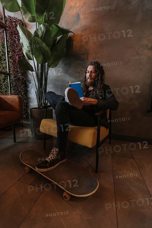 A man with long hair sits comfortably in a chair of a cafe, engrossed in a book. A skateboard rests on the floor beneath his feet, next to a large potted plant and modern decor