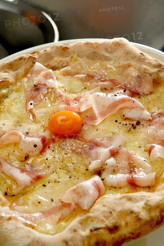 A freshly baked thin crust pizza topped with mozzarella, cured ham, and a vibrant egg yolk, seasoned with black pepper, captured up close