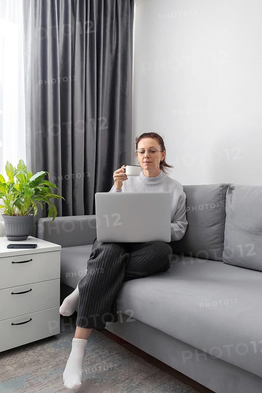 A woman work comfortably on a sofa with a laptop on her lap, sipping coffee. The scene includes gray curtains, a potted plant, and a side table, evoking a sense of calm and relaxation