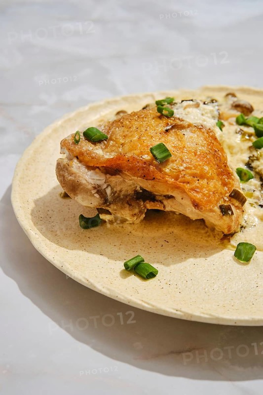 Close-up view of roasted chicken on a light-colored plate. Chopped green onions are scattered on top. The setting appears to be a kitchen with natural light coming in