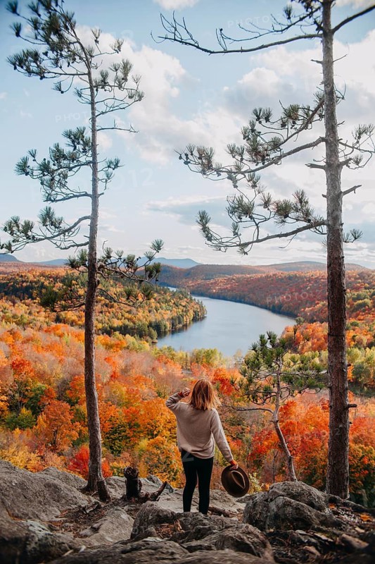 A woman stands on a hilltop, overlooking a stunning autumn landscape in Quebec, Canada. The vibrant fall foliage and serene lake below create a breathtaking view