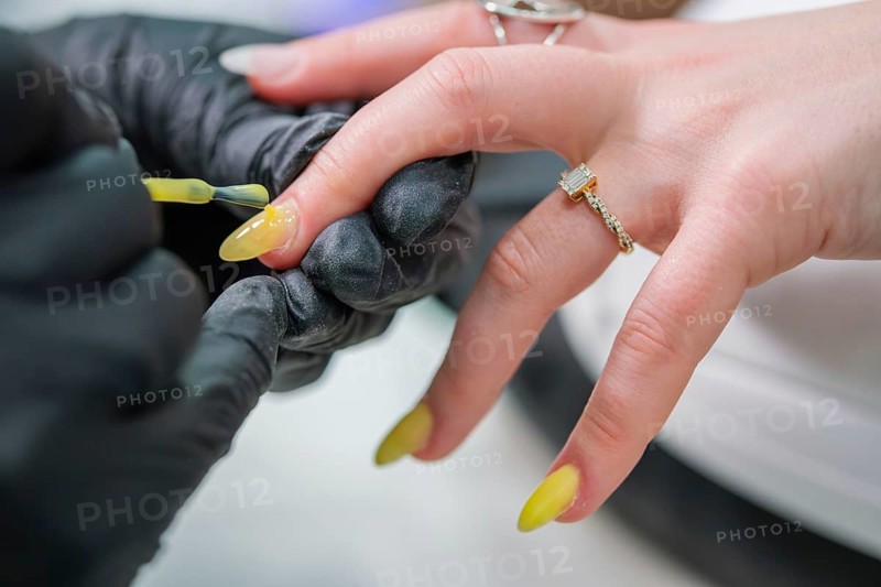 Close view of a nail technician in black gloves brushing glossy yellow polish onto a long almond nail during a professional salon manicure