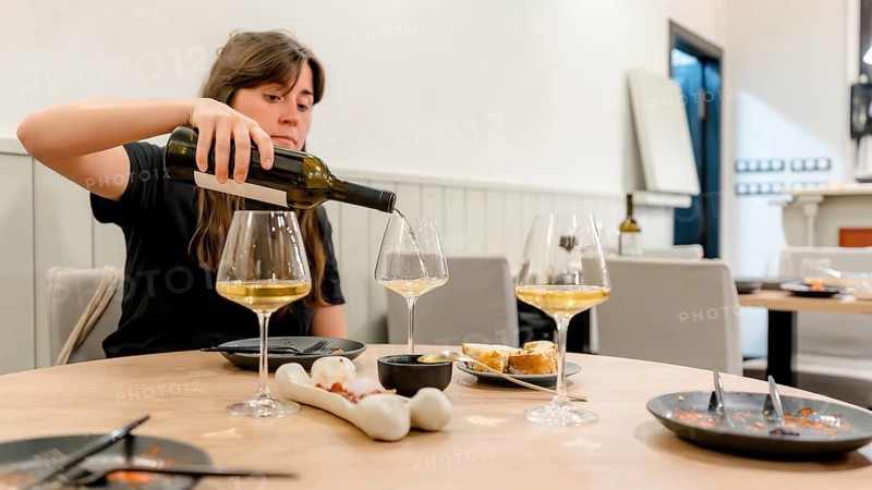 A woman pours white wine into glasses on a restaurant table set for dinner. The scene includes plates with appetizers, bread, and neatly arranged tableware, evoking a sense of cozy dining