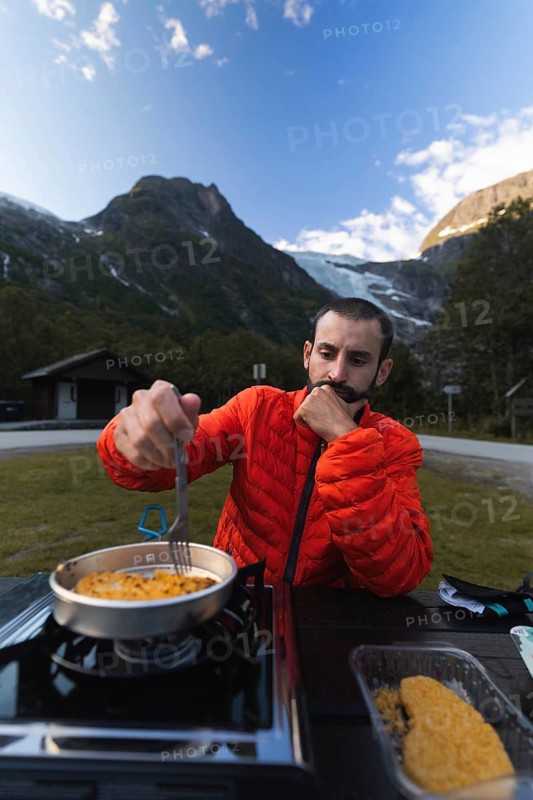 A traveler in a red jacket cooks a meal on a portable stove, set against the backdrop of majestic Norwegian mountains and a serene campsite A perfect mix of nature and adventure