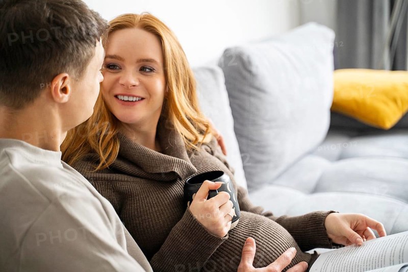 A cheerful pregnant couple sits comfortably on a modern sofa, sharing a warm moment of connection. The woman holds a cup of coffee while looking at her partner