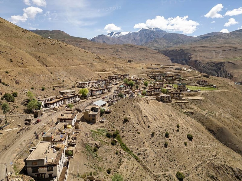 Scenic view of a tranquil village nestled in the arid mountains of Spiti Valley, India, showcasing traditional architecture against the backdrop of majestic peaks under a clear blue sky