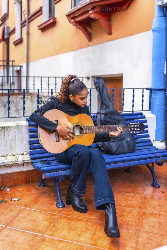 Vertical full length photo of an african american young woman playing guitar sitting on a bench outside colorful houses