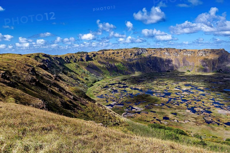 A far-reaching panorama of a crater lake surrounded by volcanic slopes under a clear sky, the Ranu Kau volcano on Easter Island in the Pacific Ocean