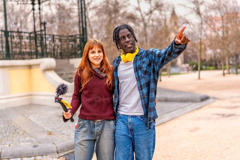Two smiling content creators are working in a park, recording audio with a boom microphone and headphones