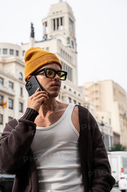A stylish man in a mustard beanie and sunglasses talks on the phone against a city backdrop. His casual outfit includes a white tank top and a brown cardigan