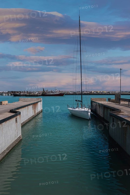 A sailboat calmly docked in a marina canal in Civitanova Marche, Italy. The sky is painted with stunning pastel colors during sunset, providing a serene backdrop