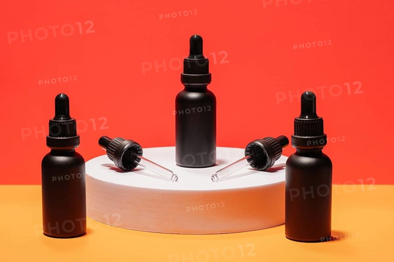 Black dropper bottles with pipettes displayed on a white platform, set against a vibrant red and orange background. Perfect for beauty and cosmetic product presentations