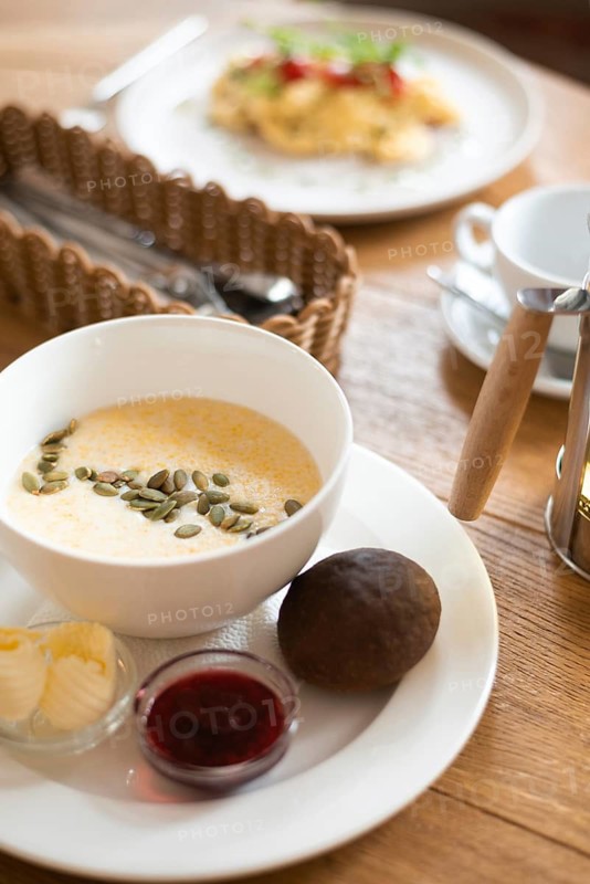 A cozy breakfast setting in a cafe featuring a bowl of creamy corn porridge garnished with seeds, accompanied by scrambled eggs, butter, jam, and a rustic roll