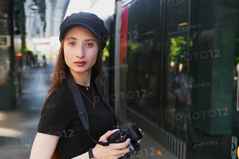 Young female photographer in urban setting, wearing casual attire, holding a camera. She stands confidently on a bustling city street, ready to capture urban scenes
