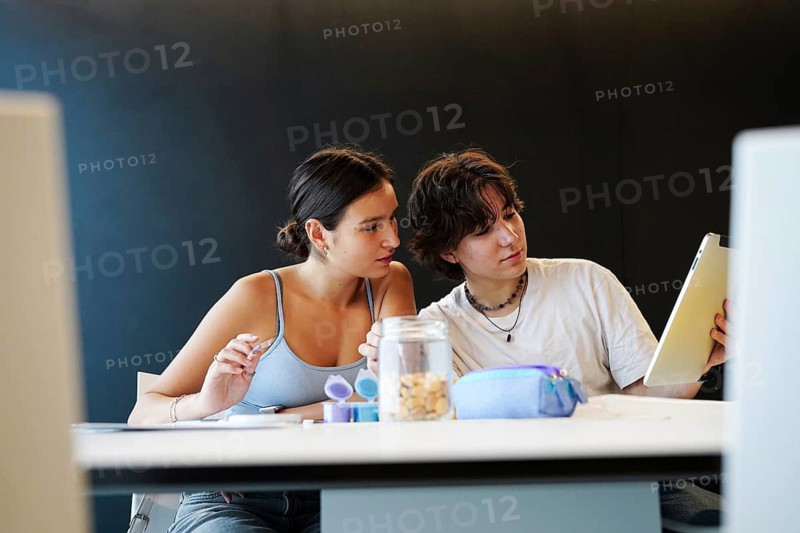 A non-binary teenager and a female student sit closely together at a school desk, engaged in shared educational activities. The non-binary individual holds a digital tablet, likely discussing educational content
