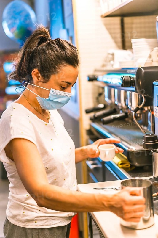 Photo session with a waitress with a face mask in a bar. New normality, security measures after the coronavirus pandemic, social distance, covid-19. Throwing milk into a cup of coffee