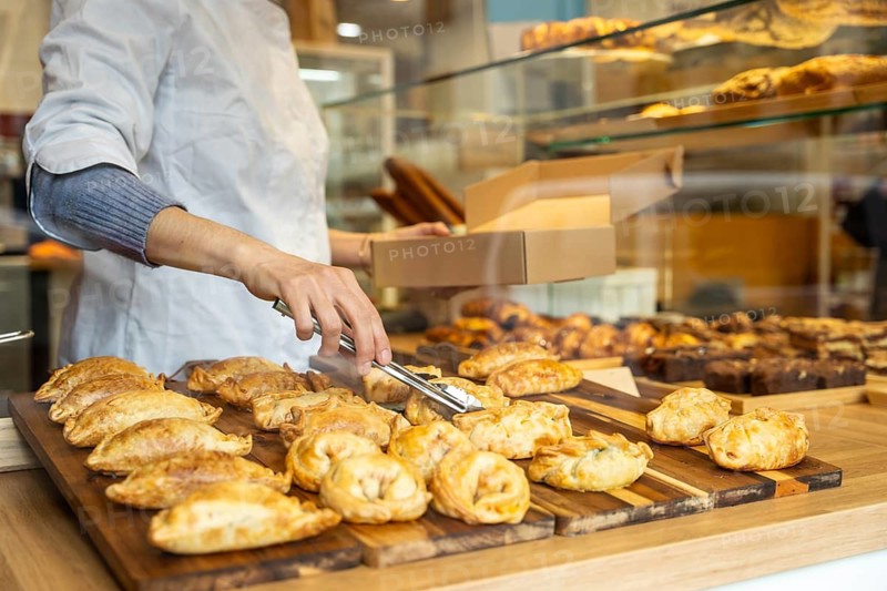 Bakery worker using tongs to place freshly baked golden savory empanadas into a brown takeaway box at the shop counter, ready for customer pickup and service