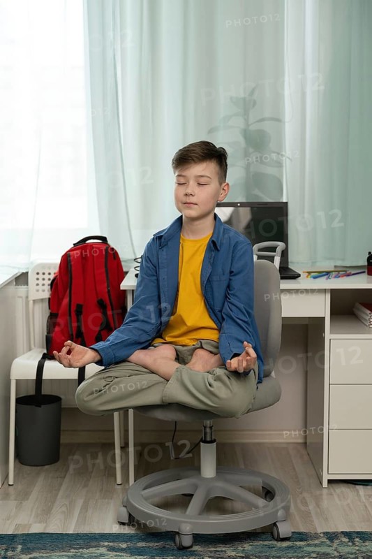A young boy sits cross-legged on a swivel chair, eyes closed, practicing meditation in a serene home setting. A desk with a laptop and colorful markers is in the background