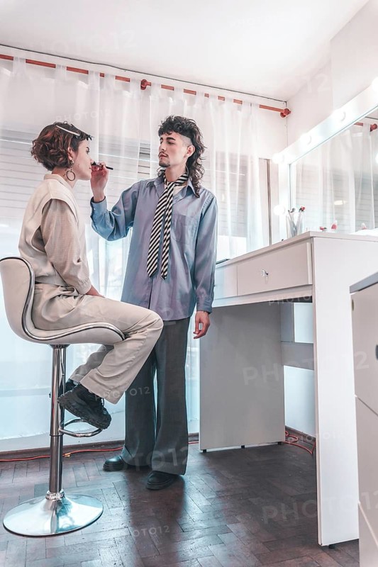 Professional makeup artist carefully applying lipstick on a model sitting on a high chair in a well-lit dressing room, preparing for a photoshoot or performance, showcasing diversity and inclusion