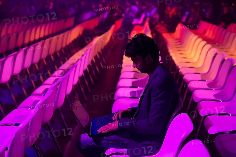 A indian man sits in a brightly lit auditorium with rows of colorful chairs at a conference of AI in Amsterdam. The atmosphere is illuminated with pink and purple hues as he focuses on his laptop