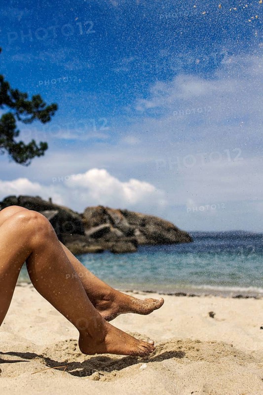 Close-up of sandy feet on a beach with a clear blue sky. The background features gentle waves and rocks, evoking a sense of relaxation and summer joy