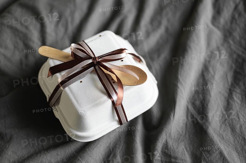 Top view of a bento birthday cake elegantly wrapped and secured with ribbon and a wooden spoon, resting on a textured gray bedsheet