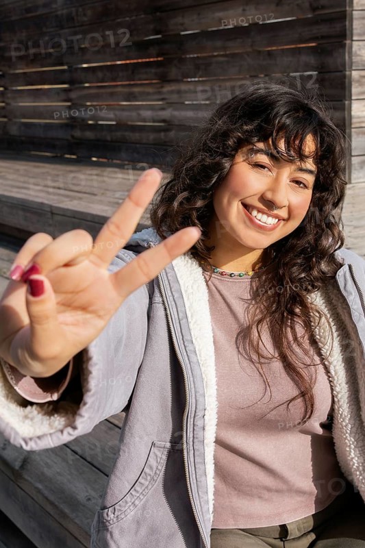 Young Latin woman smiling while enjoying a sunny day outdoors, flashing a peace sign with her fingers, expressing joy and positivity in a casual, relaxed atmosphere